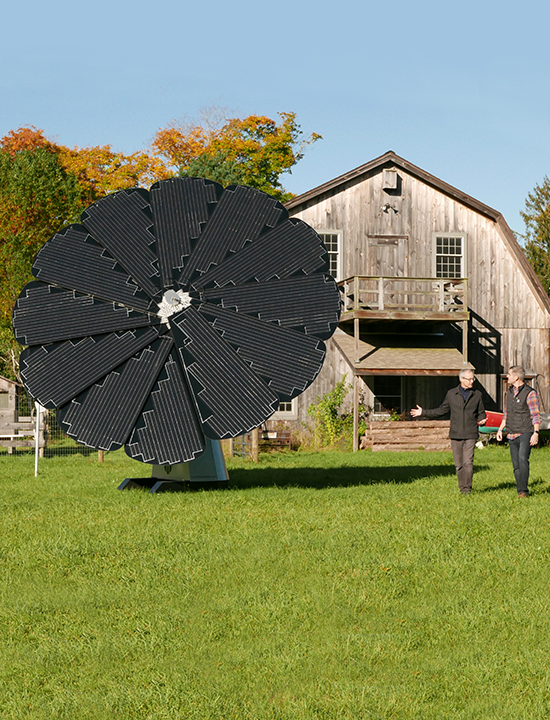 Two men walking past solar flower installation near farmhouse with autumn trees in the background